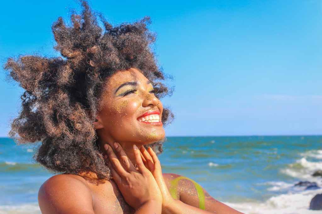 woman smiling at the beach, afro flowing in the wind, smile towards the sun, body full of glitter.